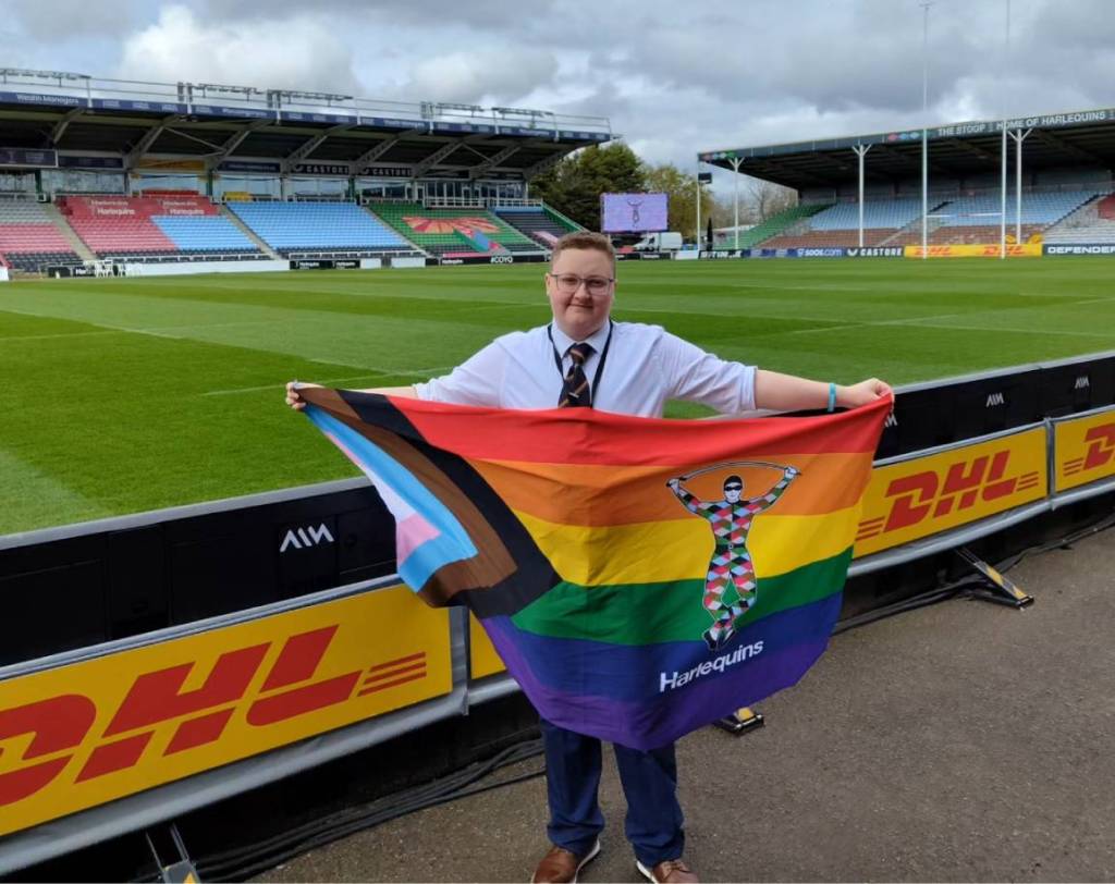 Trans man Emmett Peacock wears a white shirt and black tie as he holds up a progressive Pride flag while standing on a rugby pitch