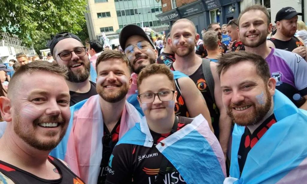 Emmett Peacock wears a rugby uniform with a trans Pride flag draped around his shoulders with other rugby players surrounding him