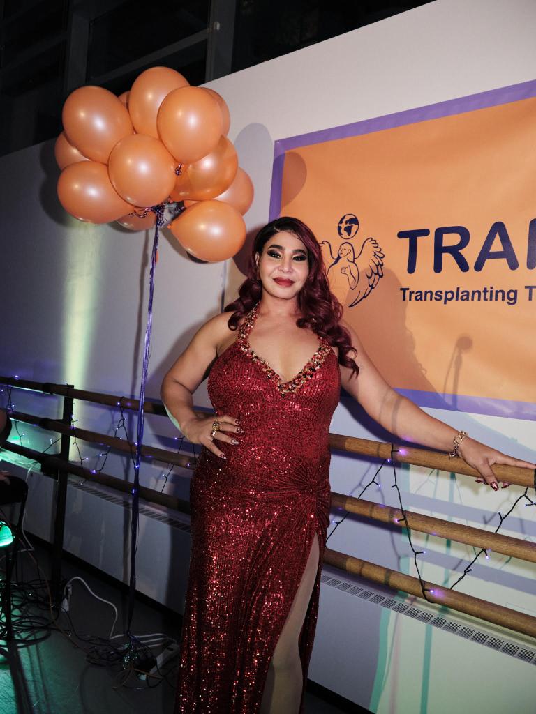 Iman Le Caire wears a red sparkly dress as she stands in front of a sign for TRANS ASYLIAS, an organisation that helps trans people find asylum abroad