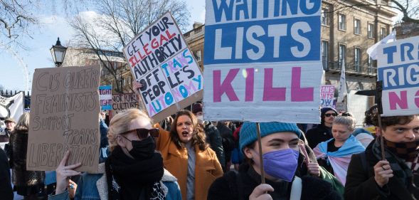 A person holds up a sign in the colours of the trans Pride flag (blue, pink and white) reading 'Waiting lists kill' referring to the huge wait times trans people face accessing gender-affirming healthcare on the NHS