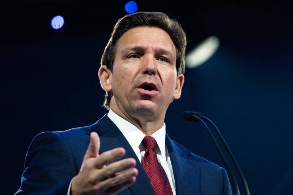 Florida governor Ron DeSantis wears a suit and tie as he gestures with one hand while speaking to people off camera