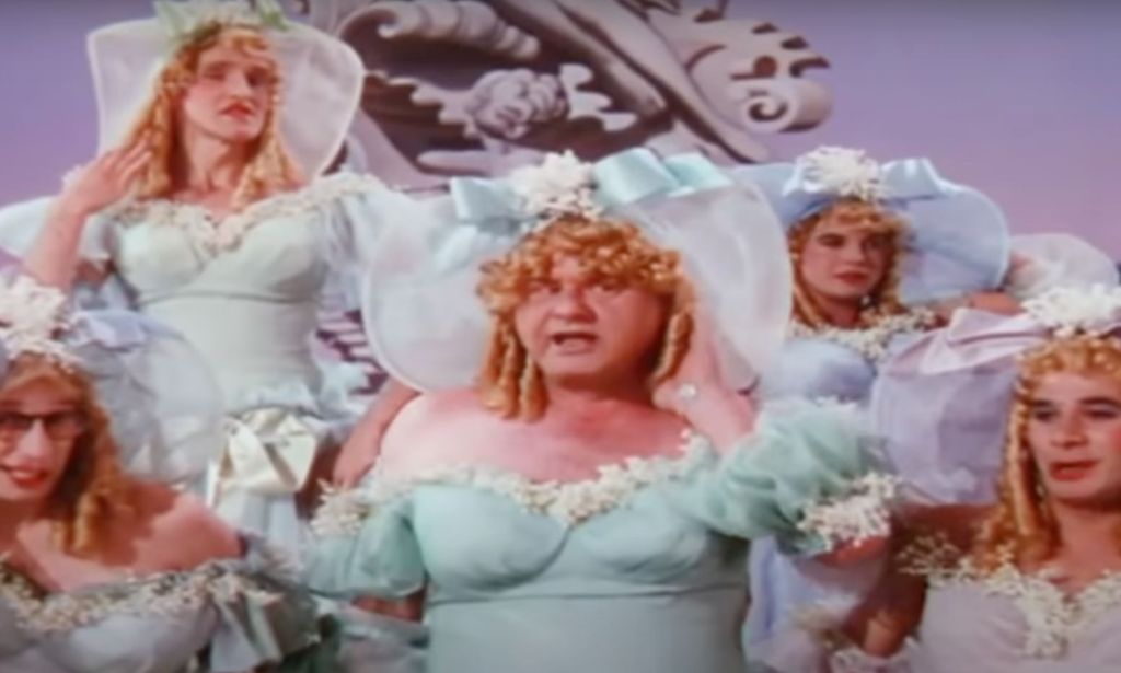 Several male actors wear blue dresses, blonde wigs and blue hats as they perform in the film This Is the Arm