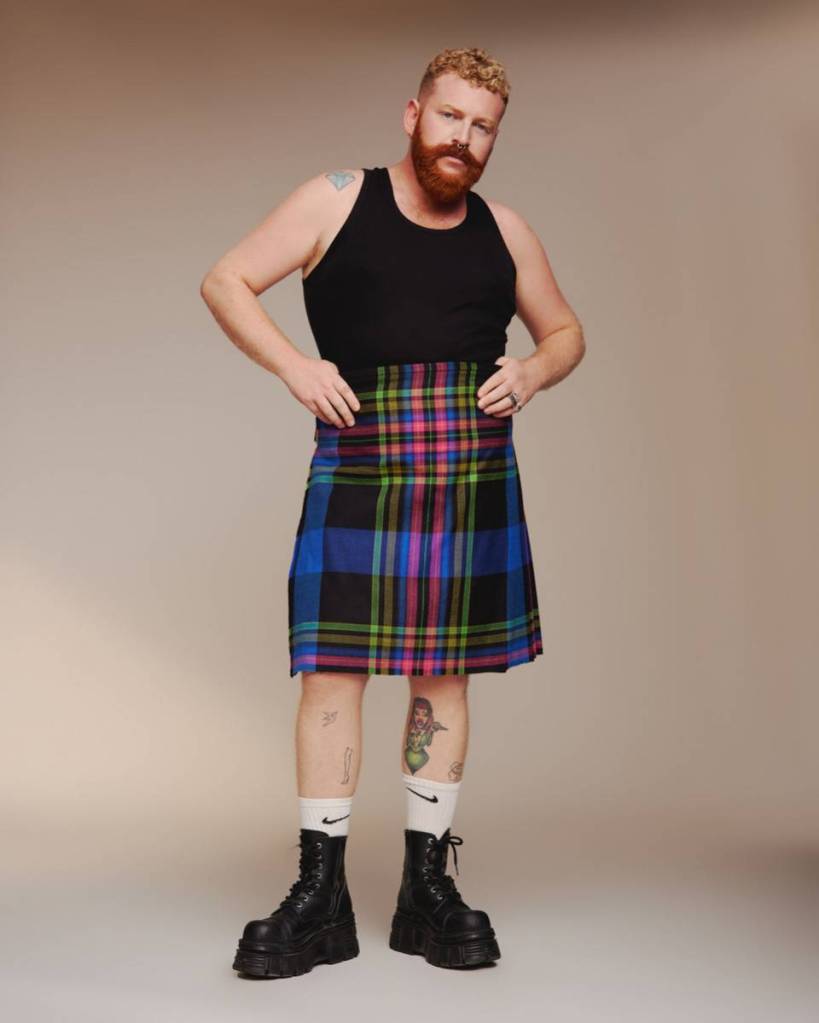 Non-binary, trans singer Tom Rasmussen wears a black sleeveless top with a rainbow plaid skirt, white socks and dark boots as they stand in front of a beige background