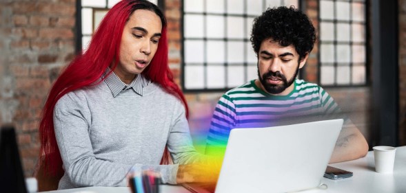 There is a feminine presenting person ont he left side of the photo. They have bright red hair and are wearing a grey shirt. Next to them is a masculine presenting person wearing a beard and a striped shirt. They are both looking down at a laptop computer that is emitting the pride flag colours.