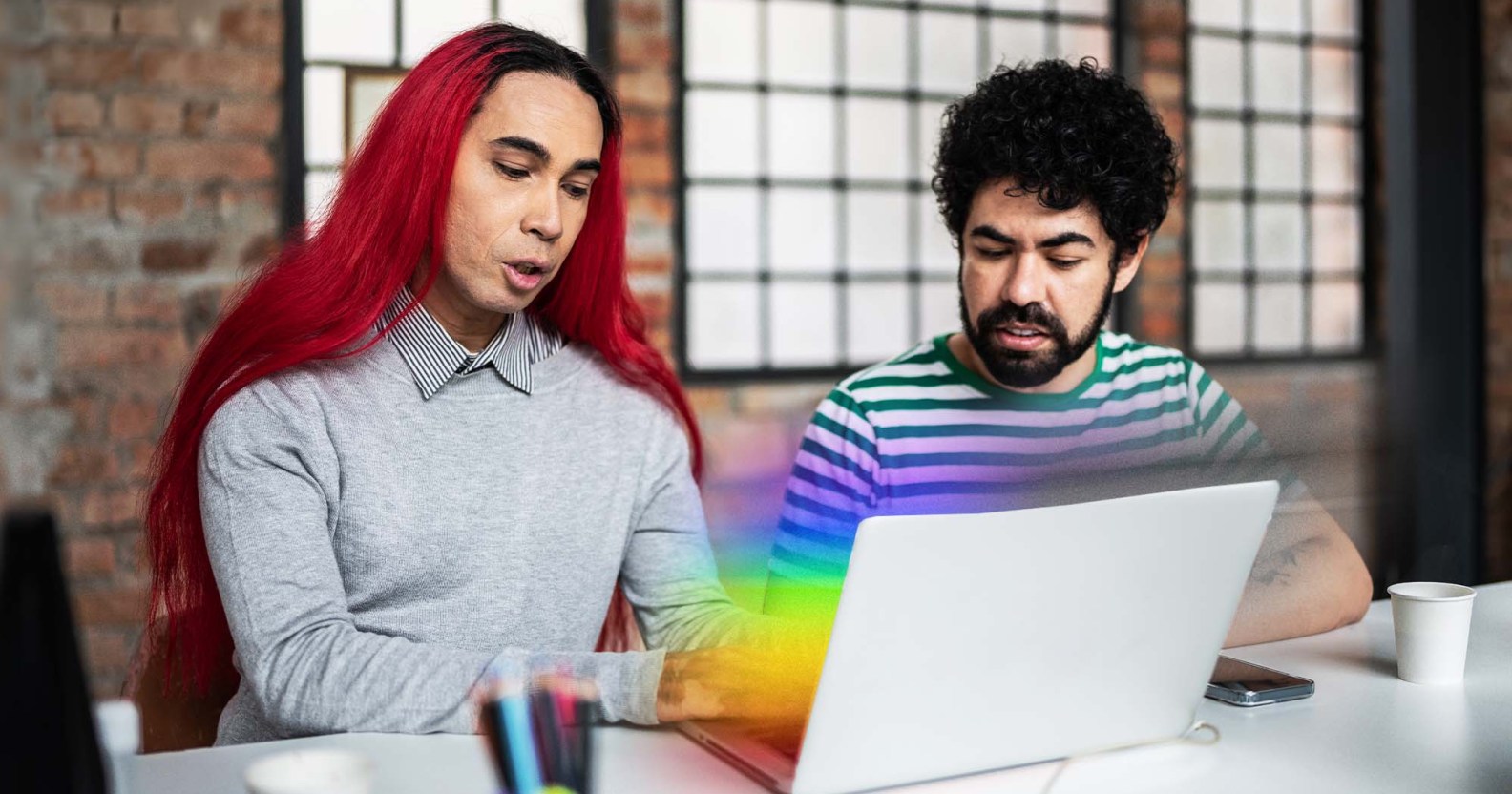 There is a feminine presenting person ont he left side of the photo. They have bright red hair and are wearing a grey shirt. Next to them is a masculine presenting person wearing a beard and a striped shirt. They are both looking down at a laptop computer that is emitting the pride flag colours.