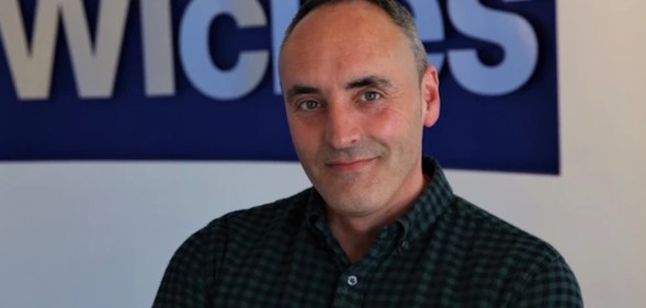 This is a portrait of Ben Jackson. He is wearing a dark shirt and has his arms folded. He is standing in front a Wickes sign.