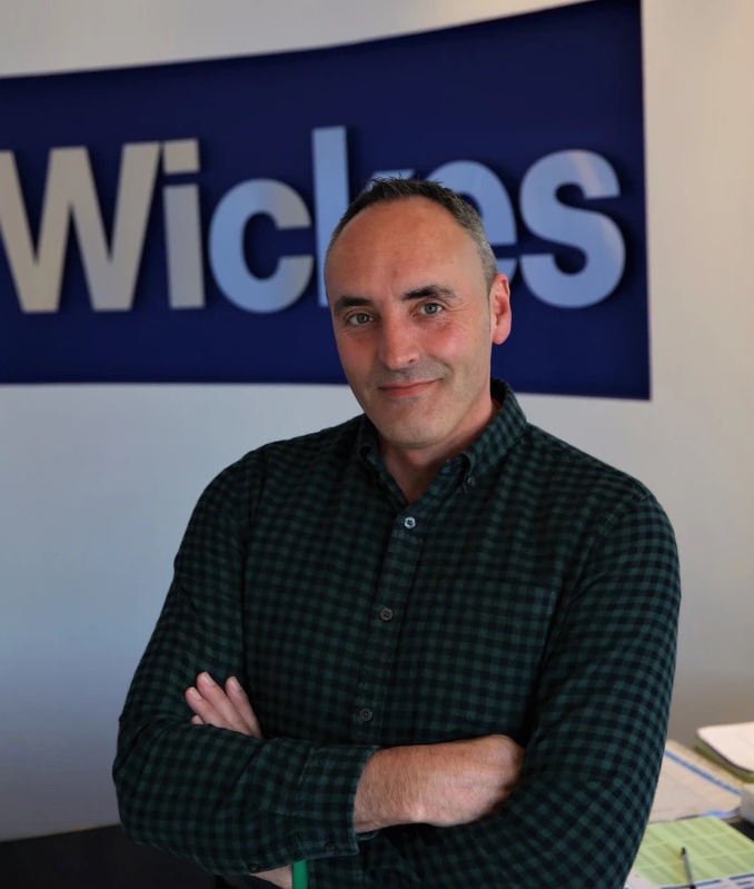 This is a portrait of Ben Jackson. He is wearing a dark shirt and has his arms folded. He is standing in front a Wickes sign.