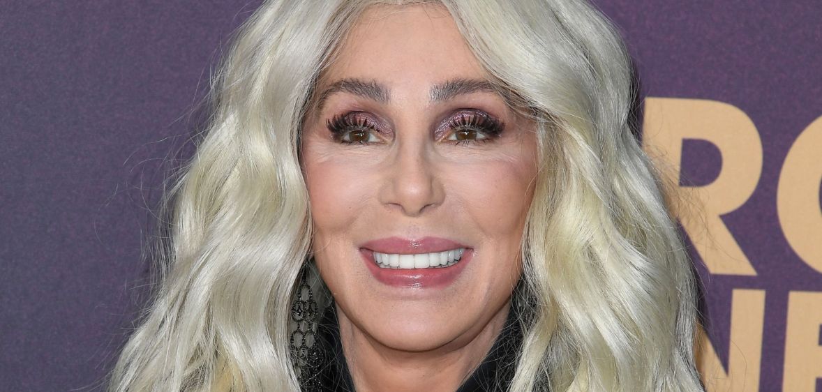 Cher smiling wearing a black coat and blonde hair, stood against a purple background with white writing on it.