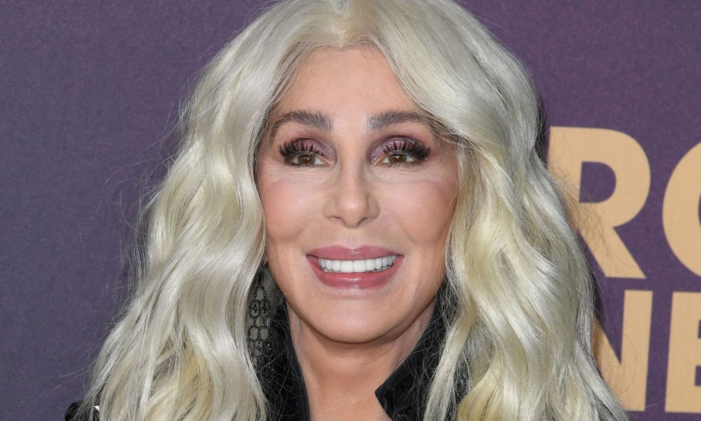 Cher smiling wearing a black coat and blonde hair, stood against a purple background with white writing on it.