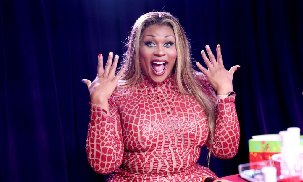 RuPaul's Drag Race icon Peppermint looks surprised and holds her hands in the air while wearing a patterned red top.