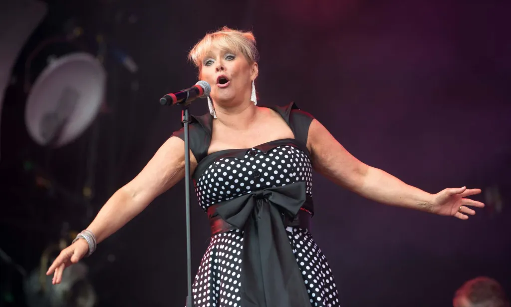 Cheryl Baker performs with a microphone, wearing a black and white polka dot dress.