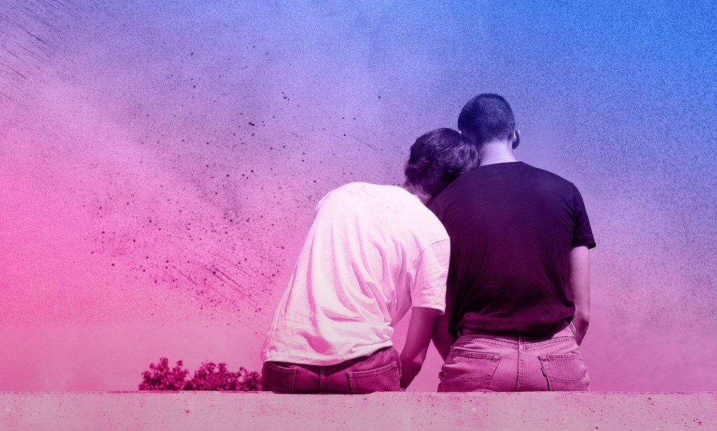A same-sex couple sitting on a wall facing away from the camera, as one rests their head on the other's shoulder