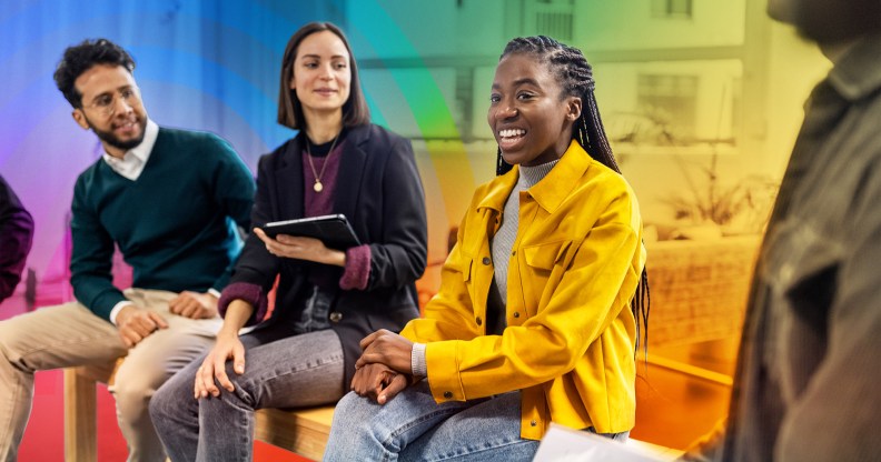 This is an image of a black woman smiling and in the middle of her colleagues. She is wearing bright yellow. The background is creatively accented with the colours of the Pride flag.