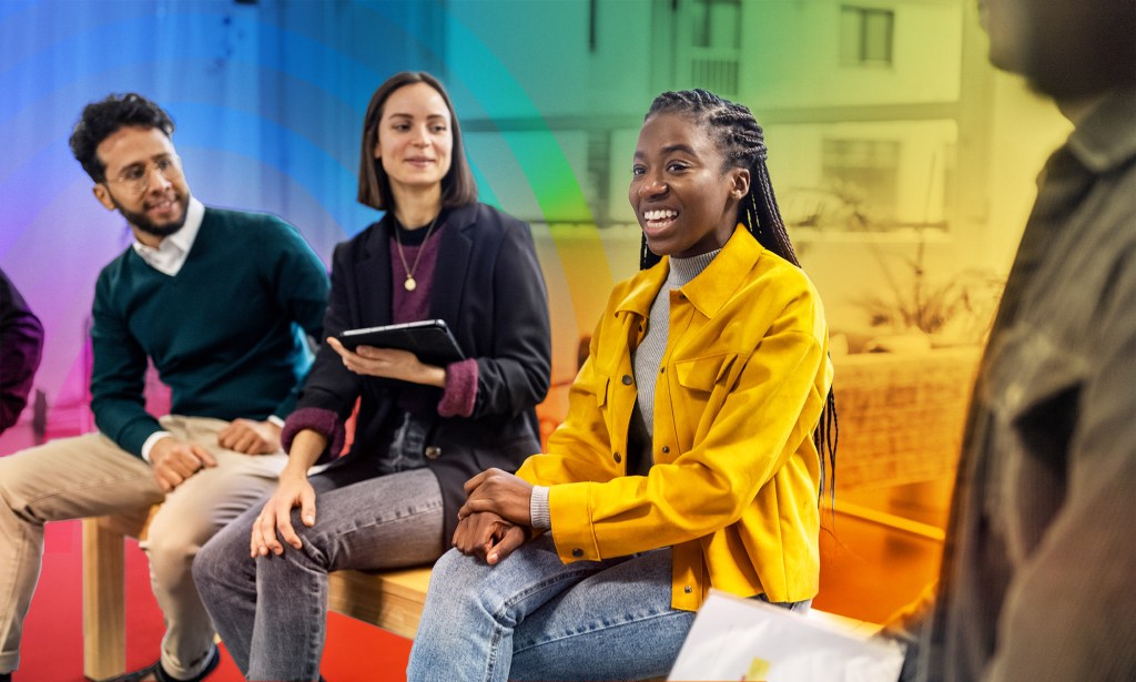 This is an image of a black woman smiling and in the middle of her colleagues. She is wearing bright yellow. The background is creatively accented with the colours of the Pride flag.
