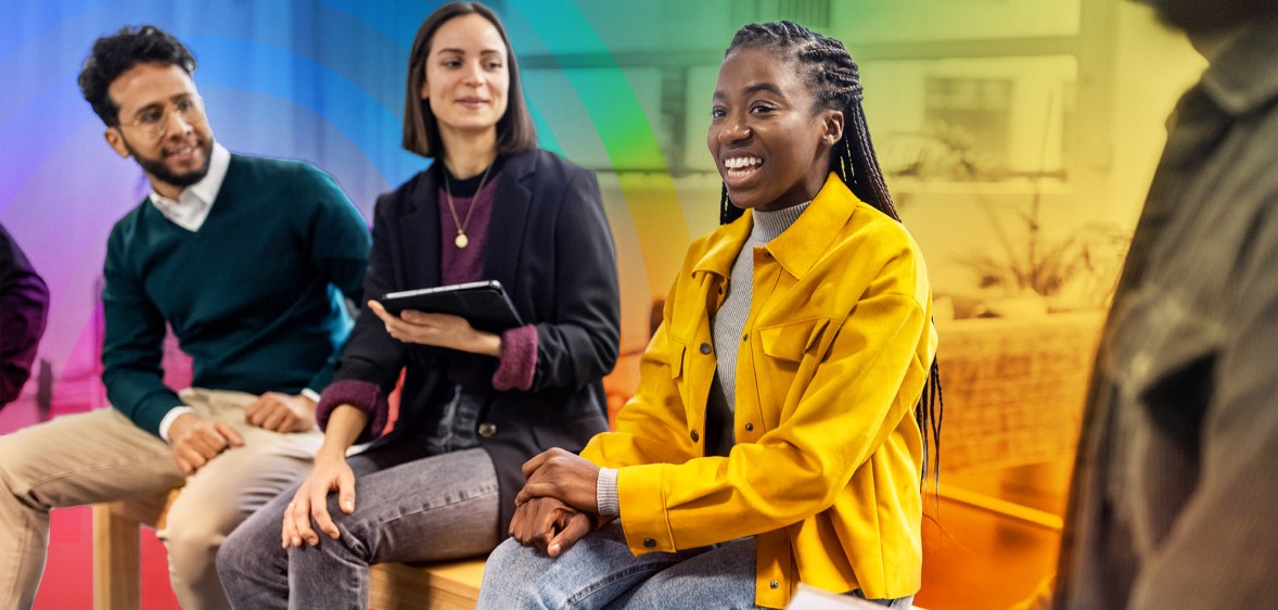 This is an image of a black woman smiling and in the middle of her colleagues. She is wearing bright yellow. The background is creatively accented with the colours of the Pride flag.