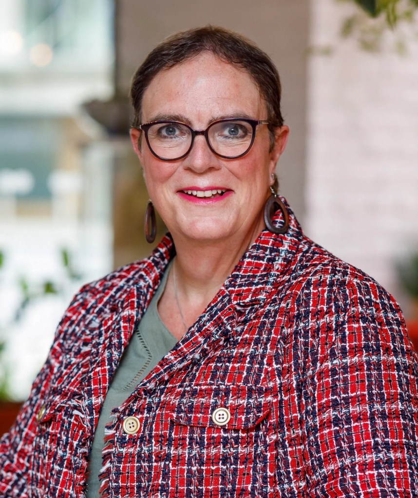 This is a professional headshot of Joanne Lockwood. She is standing and leaning on a countertop. She is wearing a plaid coat and has on dark glasses.