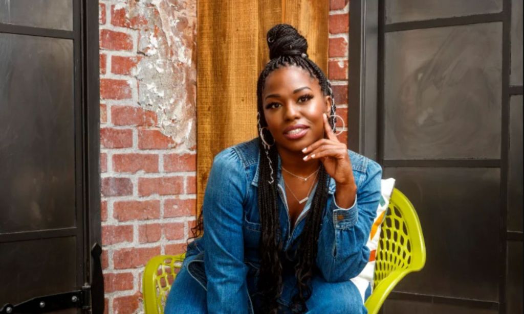 Comedian, podcast host and actress Laci Mosley plays Harper in the iCarly reboot. Here she wears a a blue jean jacket as she sits in a green chair in an exposed brick wall room.