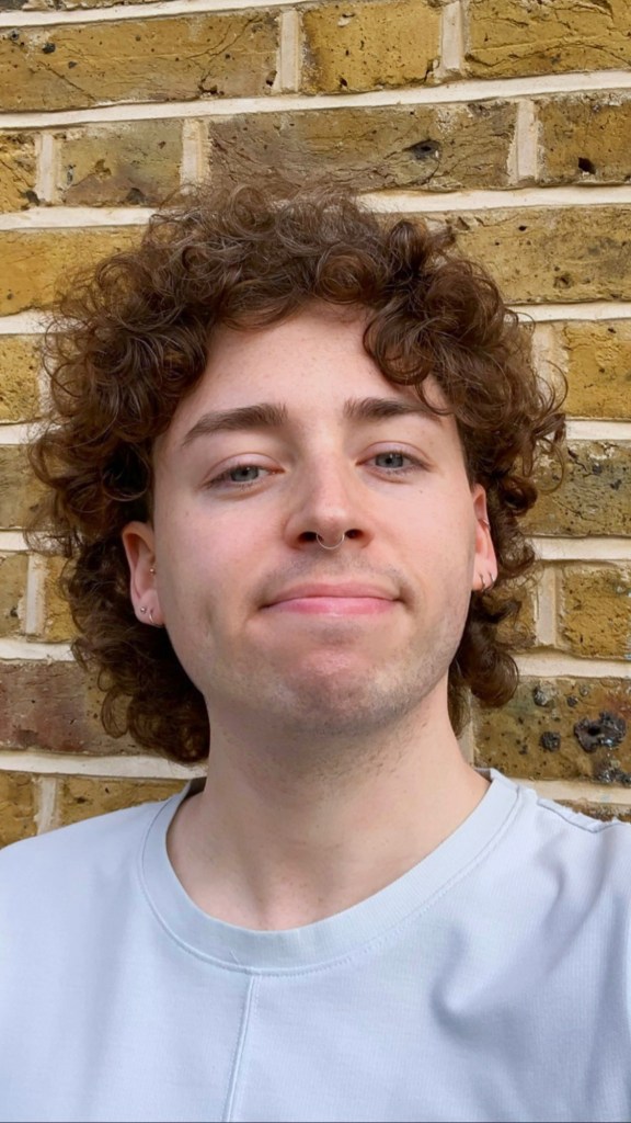 This is a headshot for Marty Davies. They are smiling and have curly brown hair. They are wearing a white shirt and standing in front of a brown brick wall. 