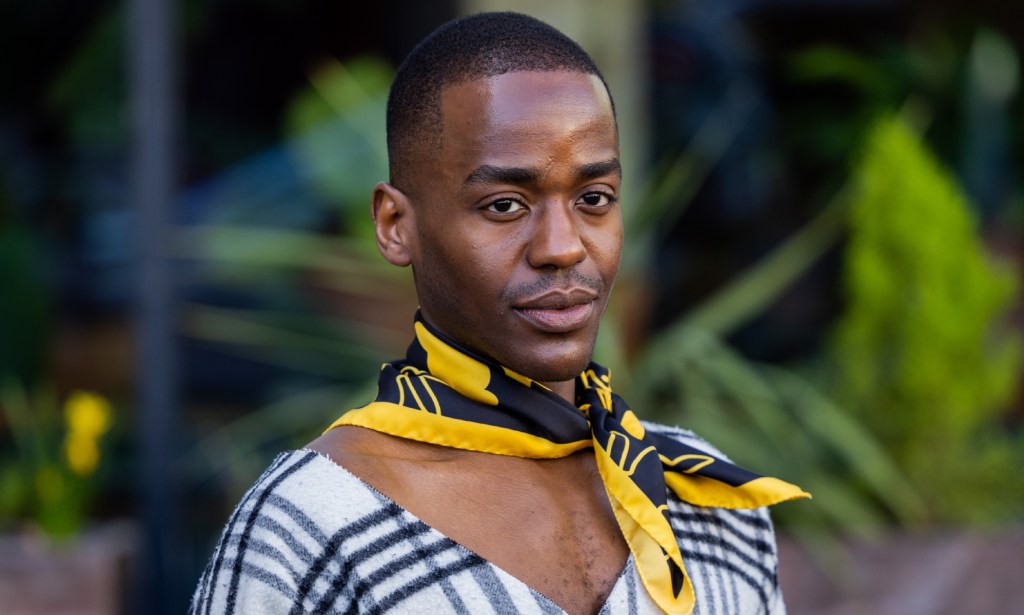 Ncuti Gatwa wearing a yellow and black necktie scarf and a white and black v-neck tartan top.