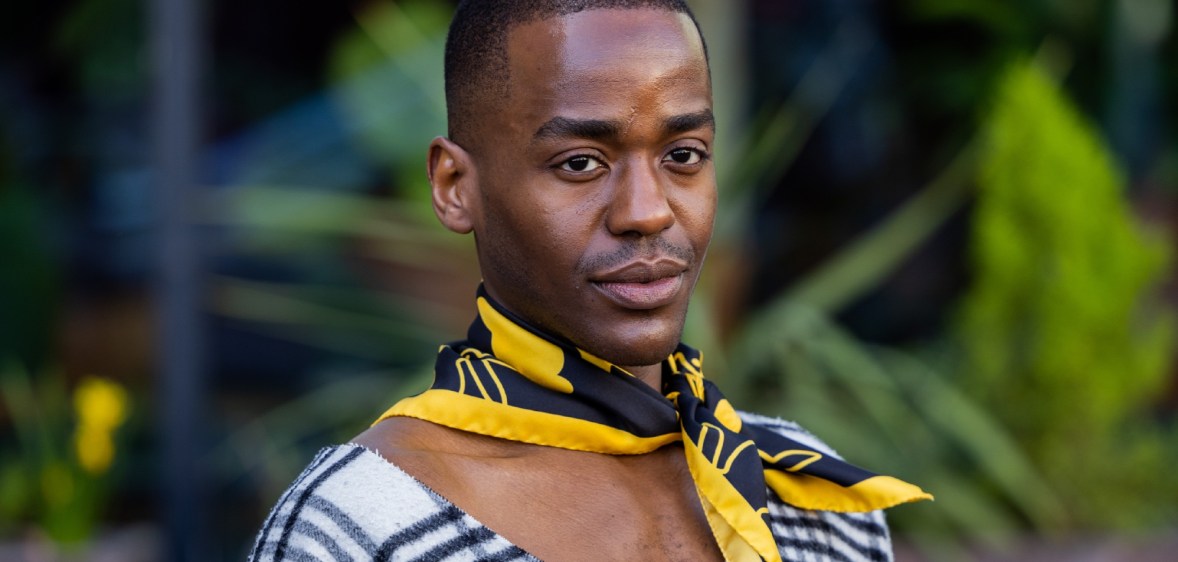 Ncuti Gatwa wearing a yellow and black necktie scarf and a white and black v-neck tartan top.
