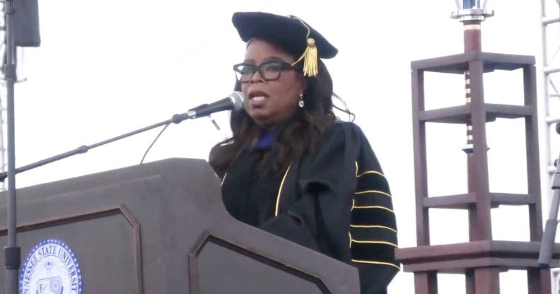 Oprah Winfrey stands behind a podium and microphone wearing a graduation outfit