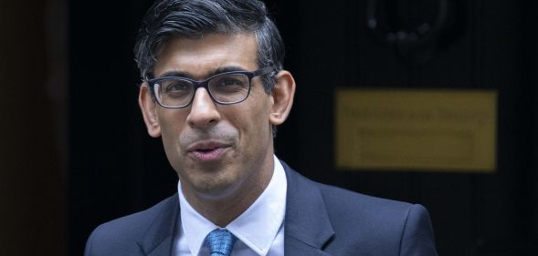 Rishi Sunak, in a suit and tie, leaves number 10 while smiling at photographers.