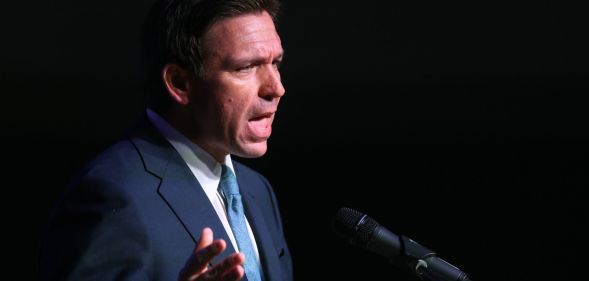 Ron DeSantis, in a blue tie and navy blue suit, speaks infront of a black background.