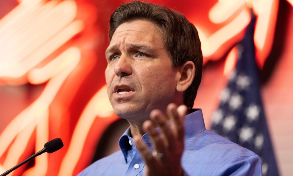 Ron DeSantis speaking to a crowd, with lights and an American flag behind him.
