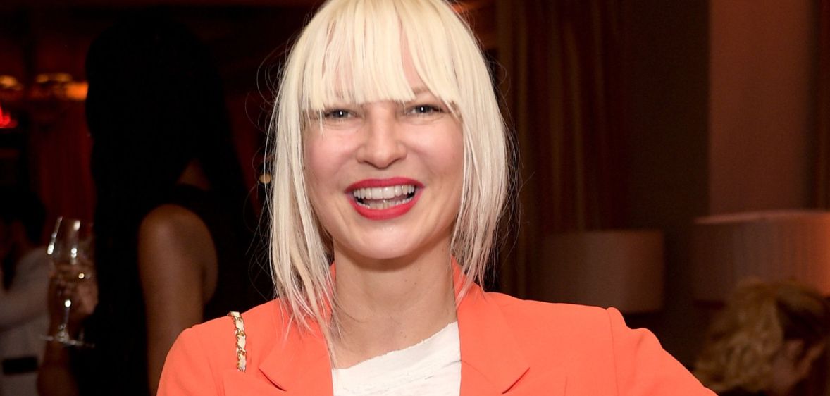 Singer Sia with blonde bangs, an orange jacket and a white top.
