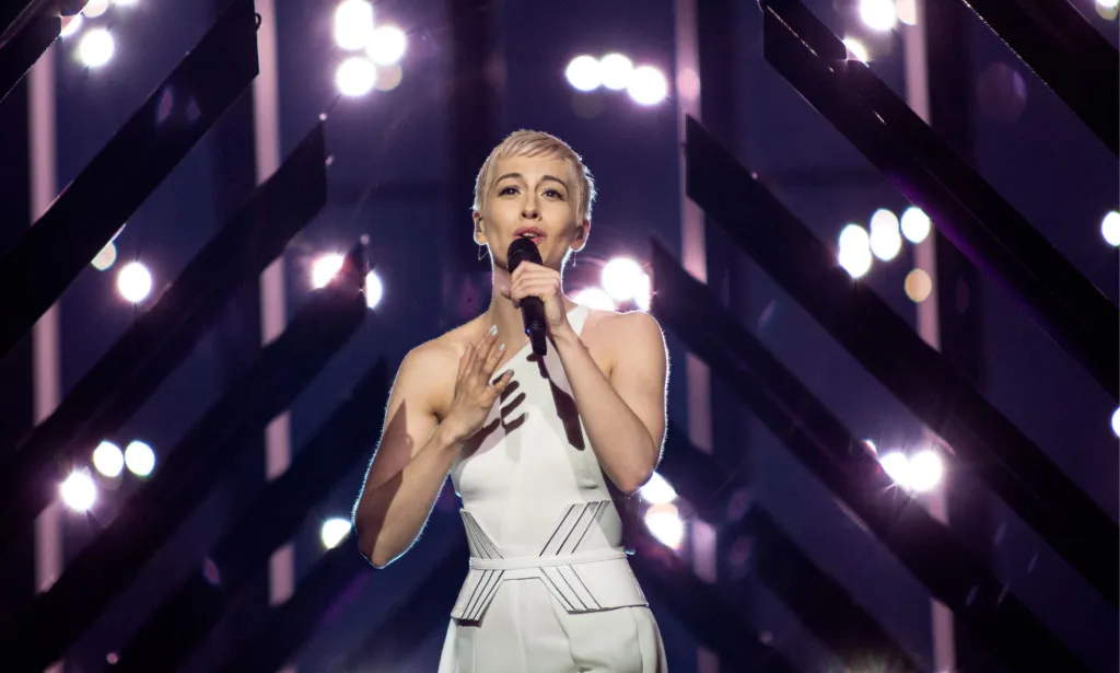 UK Eurovision act SuRie performs in a white dress in front of purple and white lights.