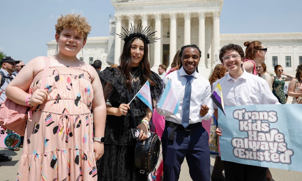 A group of four transgender youth smiling in a photo, with one of them holding a sign that reads "trans kids have always existed."