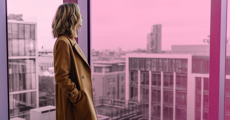 A woman is looking out of a window, they are wearing a brown coat and have blonde hair. There is a pink tint over the background which is an urban landscape.