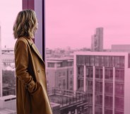 A woman is looking out of a window, they are wearing a brown coat and have blonde hair. There is a pink tint over the background which is an urban landscape.