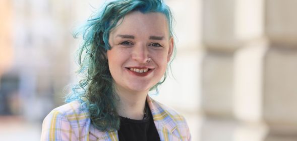 Trans activist ash Jones wears a black shirt with a purple and pink jacket on top with her hair dyed a blue-green colour