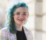 Trans activist ash Jones wears a black shirt with a purple and pink jacket on top with her hair dyed a blue-green colour