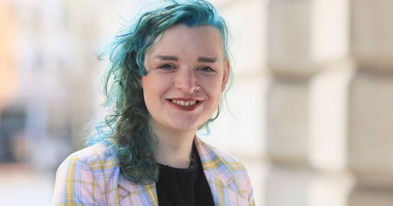 Trans activist ash Jones wears a black shirt with a purple and pink jacket on top with her hair dyed a blue-green colour