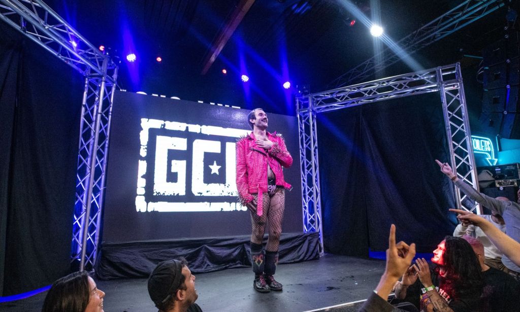 Gay wrestler EFFY wears a pink studded jacket, fishnets and black shorts as he stands before a crowd of wrestling fans