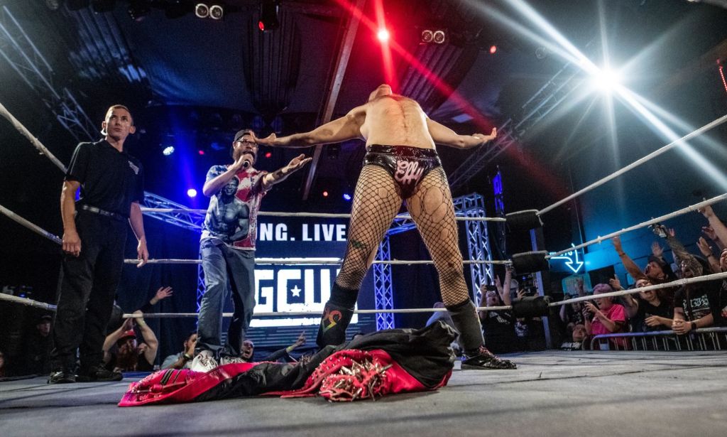 Gay wrestler EFFY stands triumphantly in the wrestling ring with his jacket on the ground in front of him as someone shouts behind him