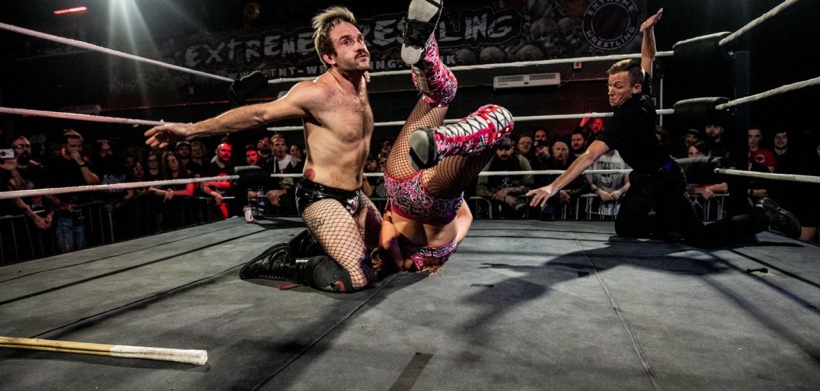 Gay wrestler EFFY kneels in the wrestling ring as he fights his opponent, who has been flipped upside down