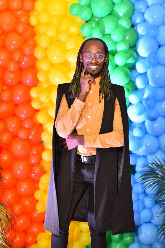Glenroy Murray, executive director of J-FLAG, wears a yellow and black outfit as he stands in front of a rainbow LGBTQ+ themed balloon background