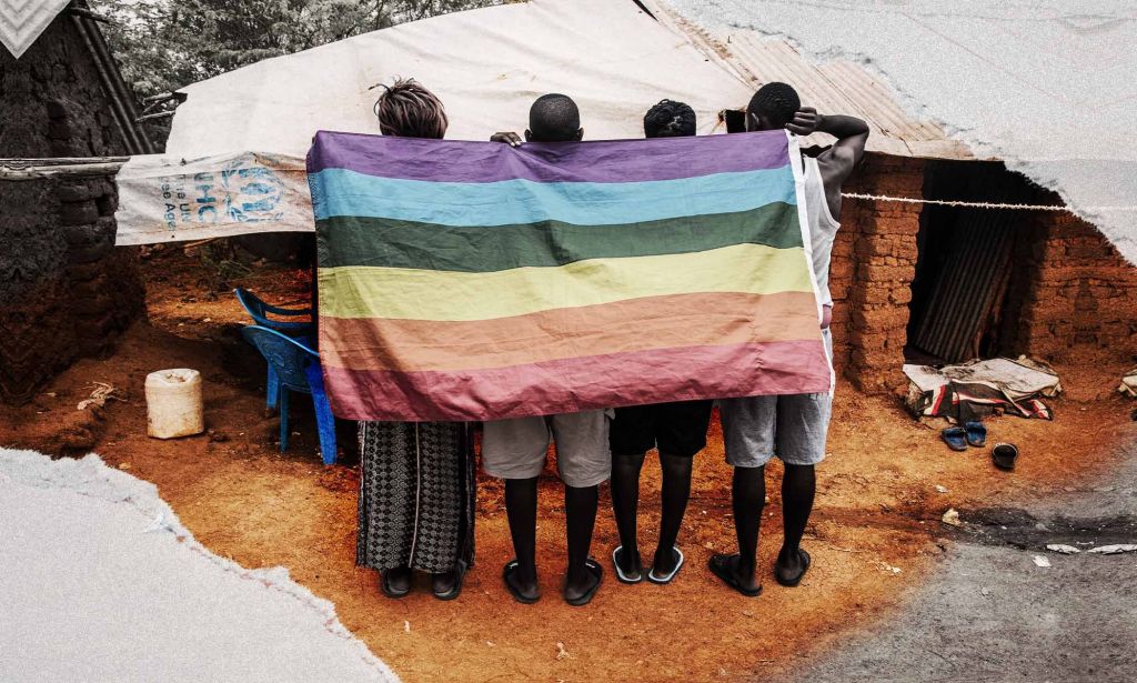 An image shows four asylum seekers standing with their backs turned to the camera in Kakuma Refugee Camp, Kenya. They are holding a Pride flag draped across their backs.
