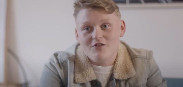 Trans teen Kai wears a white shirt and tan coloured jacket as he is interviewed for a film project by Fox Fisher and My Genderation