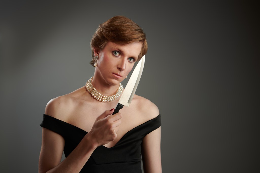 Linus Karp as Princess Diana. He is wearing a black dress and is holding a dagger in front of his face.