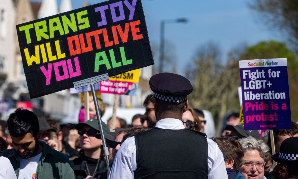A person holds up a sign reading 'Trans joy will outlive you all' in support of the LGBTQ+ community and a drag event at the Honor Oak Pub as the individual stands in front of a Met Police officer