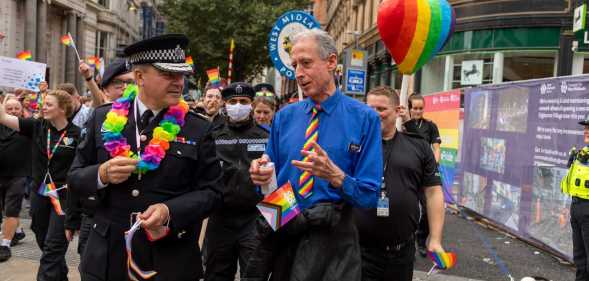 Peter Tatchell talking to the West Midlands Police chief while walking in a Pride march, surrounded by police holding rainbow flags
