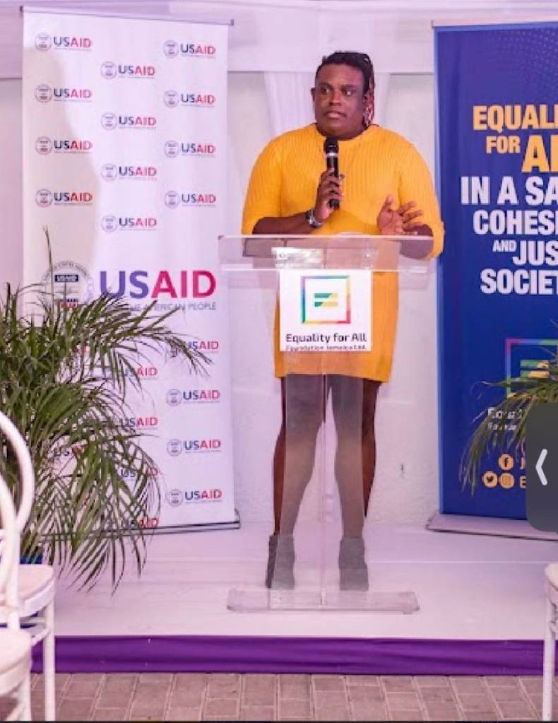 Renae Green, executive director of TransWave Jamaica, wears a yellow dress as she speaks at a podium about LGBTQ+ rights in the country