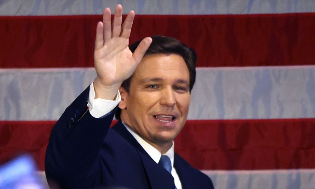 Florida governor Ron DeSantis, who is running in the 2024 Republican presidential candidate run, wears a suit and tie as he waves at a crowd off camera while standing in front of the red and white stripes of the USA flag