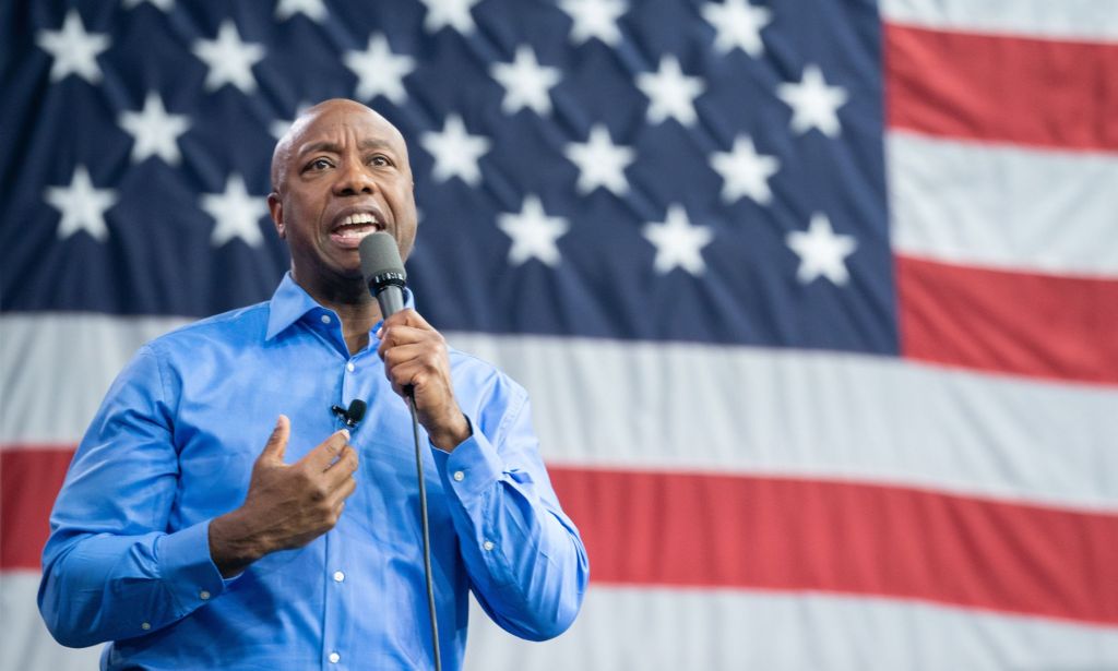South Carolina senator Tim Scott, who is a 2024 Republican presidential candidate, wears a blue shirt as he speaks into a microphone in front of a US flag