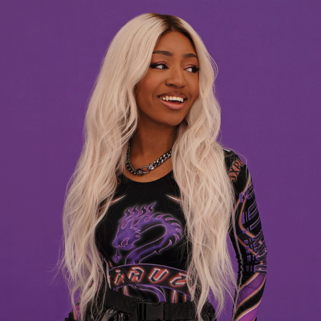 Asexual activist Yasmin Benoit wears a black shirt as she smiles and poses in front of a purple background