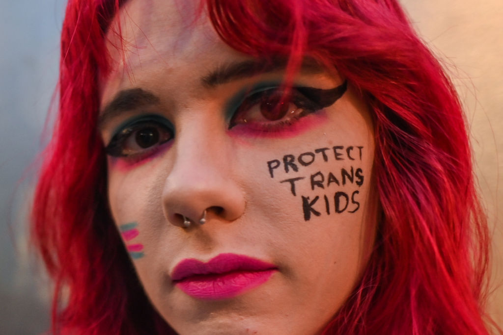 Young white person with red hair and 'protect trans kids' written on their cheek